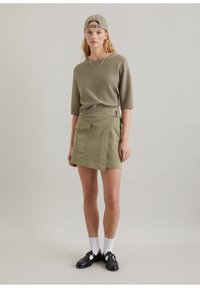Oliwkowy sweter z krótkim rękawem, zestawiony z oliwkową spódnicą typu wrap z boczną kieszenią i detalem paska. Czarne buty dopełniają całość.