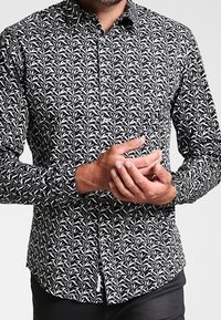 Czarna i biała koszula na guziki z kwiatowym wzorem, długimi rękawami i ostrym kołnierzykiem. Tkanina wydaje się lekka i gładka.