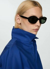 Black oval sunglasses, blue jacket with a high collar; smooth texture, button details, and a sleek design. Model has straight blonde hair.