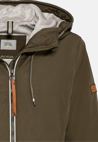 Veste à capuche vert olive avec intérieur doublé en polaire, dotée d'une fermeture éclair frontale, de détails en cuir marron et d'un patch logo sur la manche.