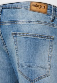 Lys blå denimjeans har et merke med "INDICODE EST. 1993" sydd på livet og en enkelt baklomme med oransje søm.
