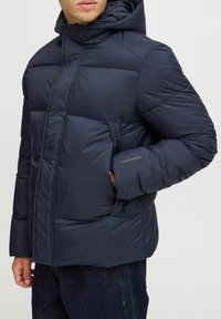 Mørkeblå puffjakke med polstret design, der har hætte, forlommer og et mærke på ærmet. Glat tekstur.