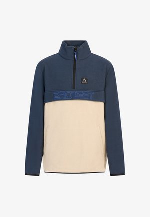 Fleece jack in marineblauw en taupe met ritskraag, voorzien van een blauw logo. Blauwe gebreide beanie. Lichtblauwe spijkerbroek en grijze sneakers maken de look compleet.