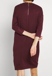 Persona che indossa un vestito bordeaux lungo fino al ginocchio, con maniche a tre quarti e cerniera visibile sul retro, in piedi con le mani incrociate dietro.