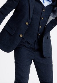 Ciemnoniebieski garnitur z teksturowanej tkaniny, z klapą w kształcie notcha, mosiężnymi guzikami oraz dopasowaną kamizelką, w połączeniu z elegancko skrojonymi spodniami.