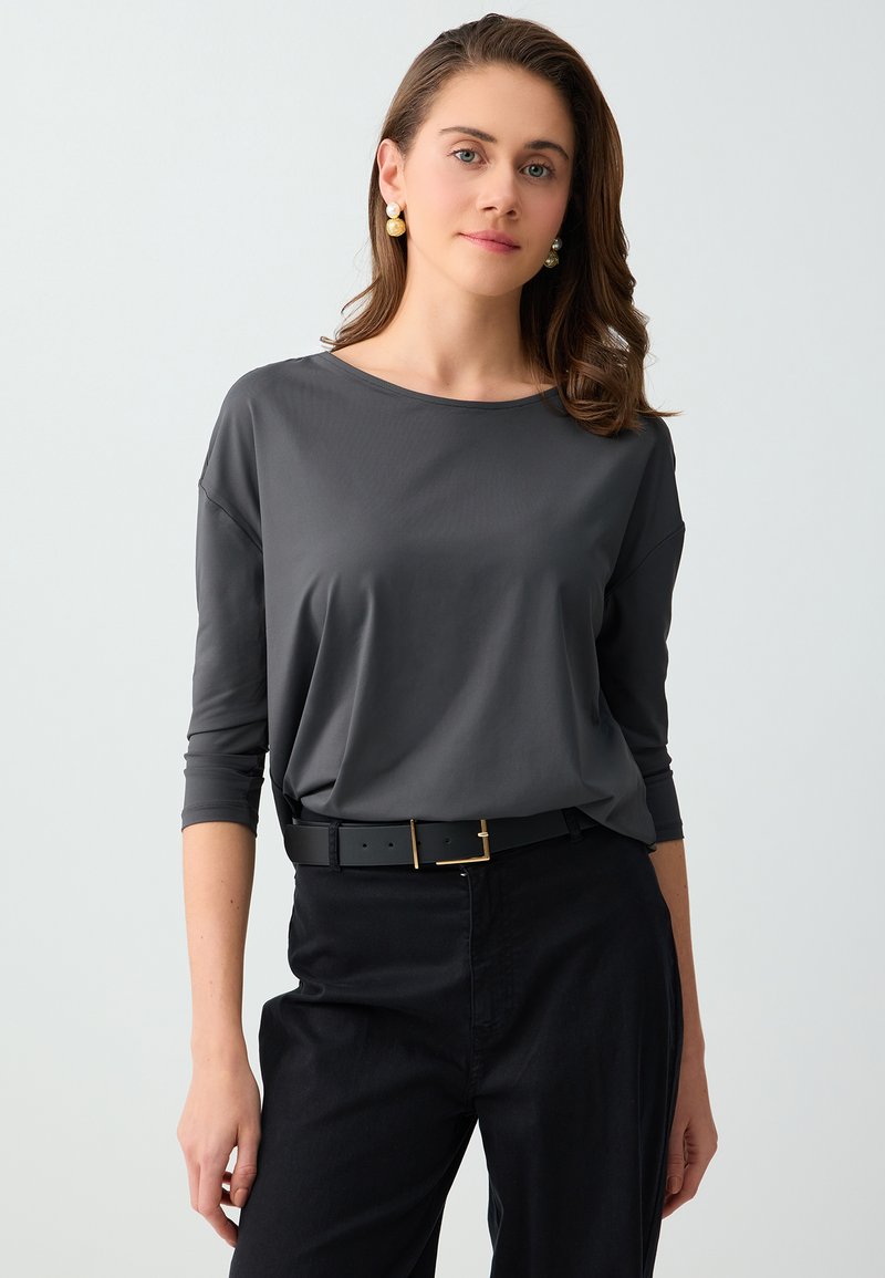 Jeune femme aux cheveux bruns ondulés porte une chemise grise à manches longues, un pantalon noir avec ceinture et des boucles d'oreilles pendantes en perle, debout devant un fond uni.