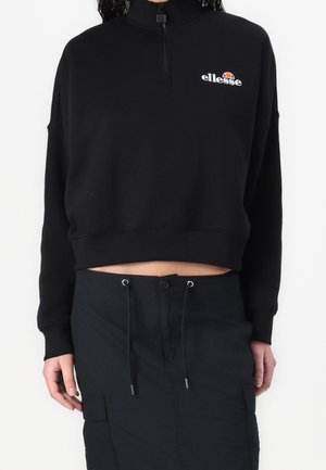 Osoba nosząca czarną, przyciętą bluzę zapinaną na zamek z białym logo „ellesse” oraz czarną spódnicę z troczkami na białym tle.