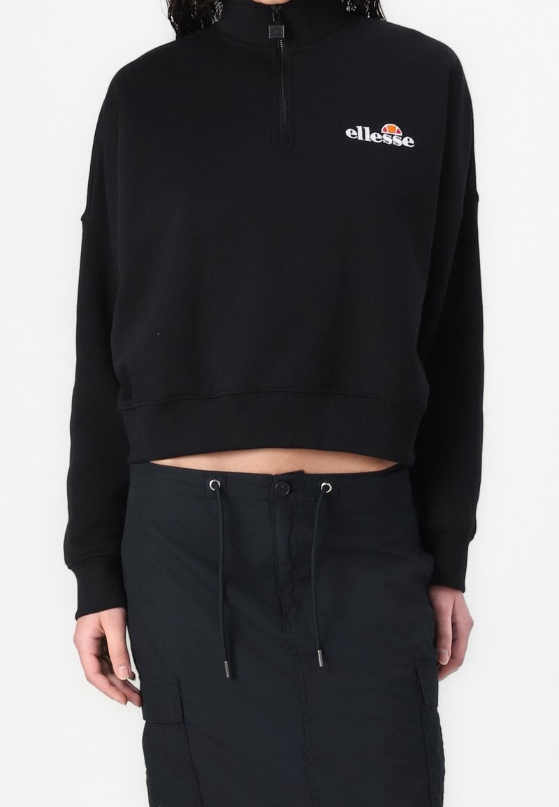 Personne portant un sweat à capuche court zippé noir avec le logo blanc « ellesse » et une jupe noire à cordon sur fond blanc.