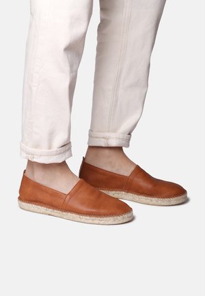 Zapatos de cuero marrón claro tipo slip-on con punta redondeada y suela de yute cosida. Lleva puestos unos pantalones crema con puños, que muestran una textura suave.