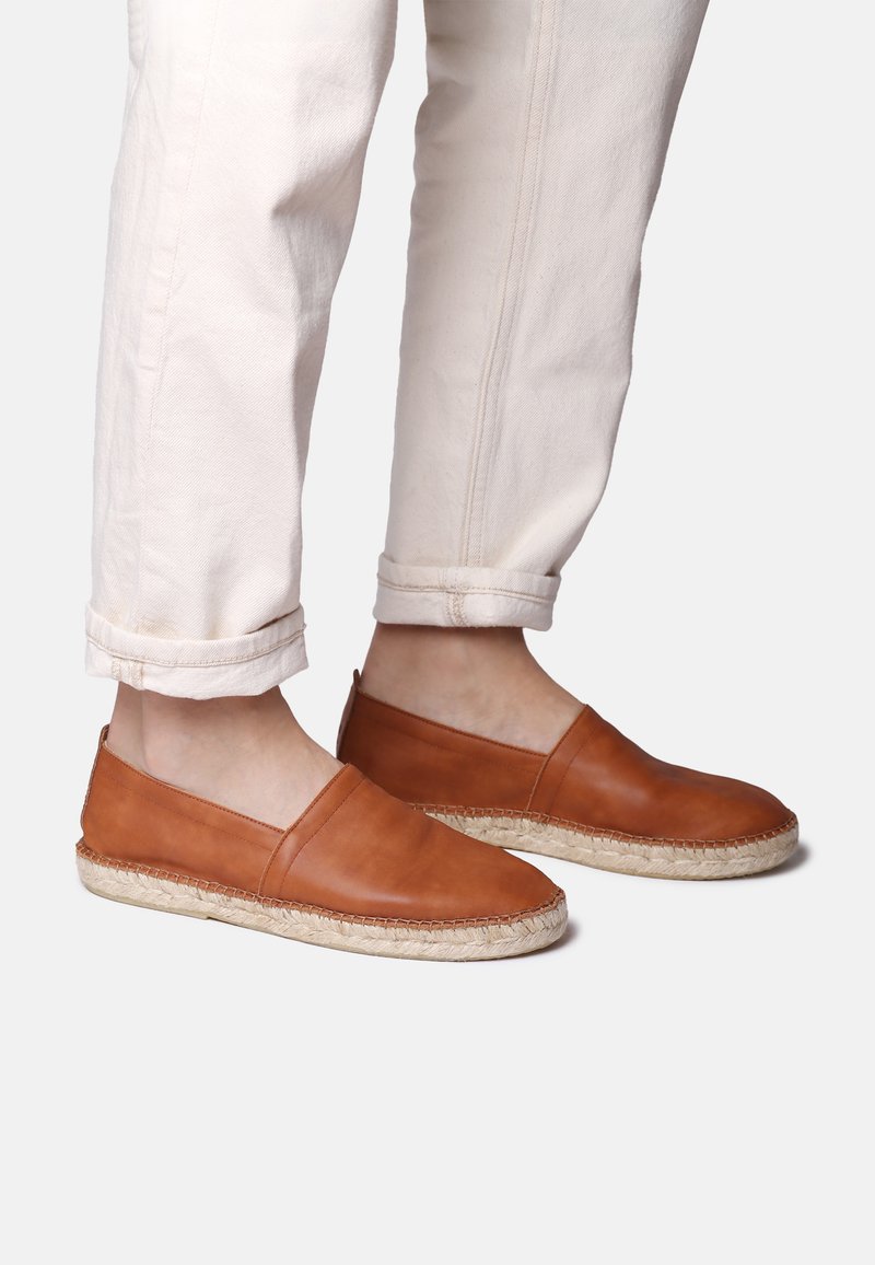 Zapatos de cuero marrón claro tipo slip-on con punta redondeada y suela de yute cosida. Lleva puestos unos pantalones crema con puños, que muestran una textura suave.