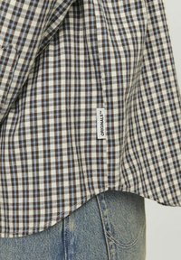 Camisa de cuadros marrón y azul con dobladillo redondeado, hecha de tela tejida, que presenta una etiqueta "ORIGINALS" visible en el lateral.