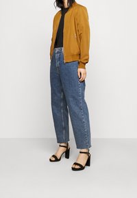 Brązowa kurtka bombowca, czarna koszula, niebieskie dżinsy z wysokim stanem oraz czarne sandały na grubej podeszwie. Gładki materiał i luźny krój spodni.