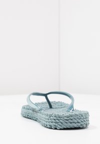 Turkos flip-flop med en texturerad vävd sula, som har glänsande, flexibla remmar och en platt profil för komfort.