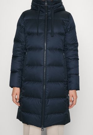 Kobieta w długim, czarnym, ocieplanym płaszczu zimowym z przednim suwakiem, zasuwanymi bocznymi kieszeniami i kapturem ze ściągaczami.