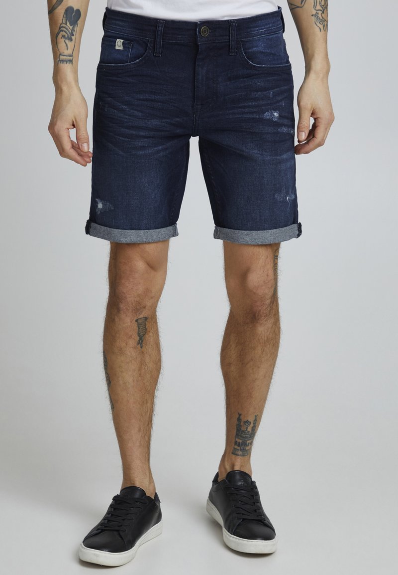 Shorts en denim bleu foncé avec une finition usée, des revers roulés et un design à cinq poches. Portés avec des baskets noires.