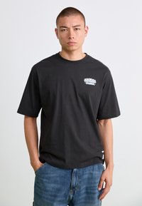 Mand med hænderne i lommerne iført sort Ripndip Puma T-shirt og blå jeans mod en ensfarvet hvid baggrund.