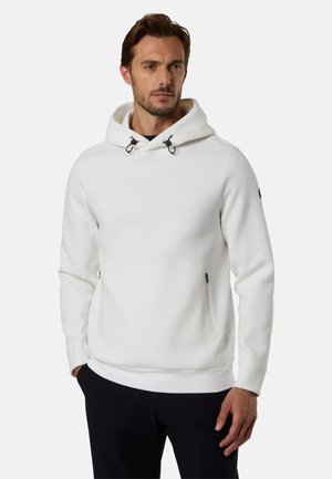 Uomo che indossa una felpa bianca con cappuccio e tasche con zip e pantaloni neri, in piedi davanti a uno sfondo chiaro e uniforme.