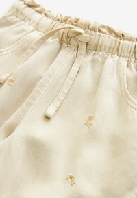 Pantaloni din bumbac bej cu talie elastică și șnur, prezentând broderii florale subtile și buzunare laterale. Textură netedă.