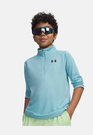 Berniukas dėvi atspindinčius mėlynus sportinius saulės akinius, šviesiai mėlyną Under Armour puloverį su ketvirčio užtrauktuku ir šviesiai žalius šortus su rankomis kišenėse.