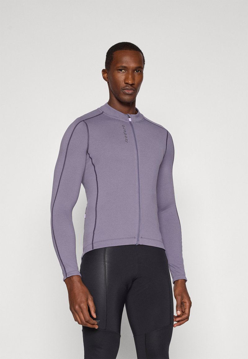 Chaqueta deportiva púrpura con cuello alto, mangas largas, cremallera frontal y costuras en contraste. Combinada con pantalones deportivos negros ajustados.