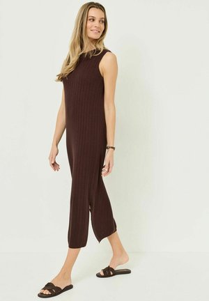 Žena kráčí, má na sobě hnědé midi šaty bez rukávů s žebrovanou texturou, s bočními rozparky a tmavě hnědé ploché sandály, na světlém pozadí.