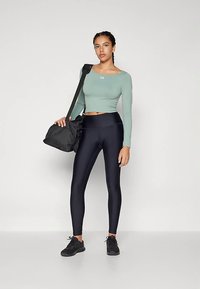 Top crop cu mâneci lungi de culoare verde deschis, colanți negri cu talie înaltă și finisaj lucios, și adidași negri. Modelul poartă o geantă de sală neagră.