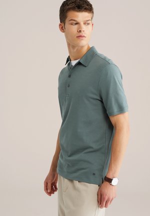 Polo met korte mouwen in teal stof met een klassieke kraag, een knoopsluiting met drie knopen en een zijschuif; gecombineerd met lichtbeige broek en een horloge.
