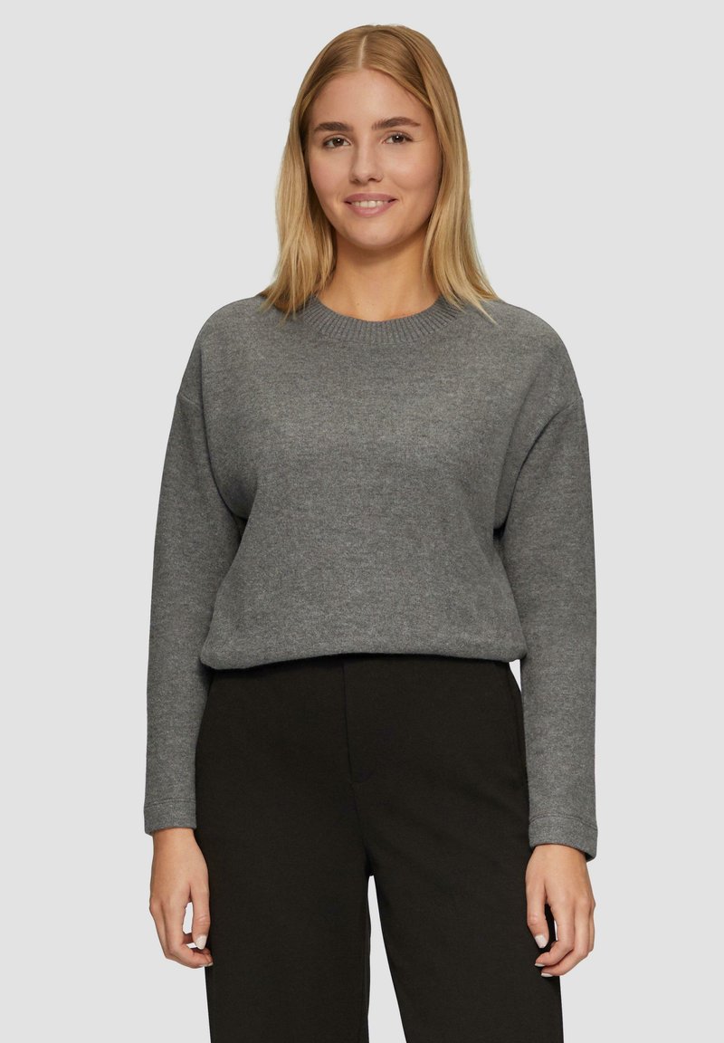 Pull gris avec un col rond, des épaules tombantes et des manches longues. Le tissu semble doux avec une coupe légèrement ample. Associé à un pantalon sombre.