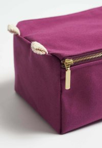 Lila tygväska med rektangulär form, gulddragkedja och krämfärgade repdetaljer på hörnen. Slät textur.