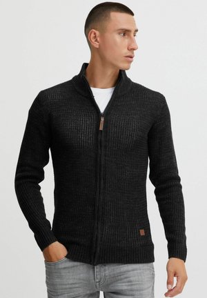 Sweat à zip noir avec une texture côtelée, un col haut et une petite étiquette en cuir brun sur l'ourlet. Porté par-dessus un t-shirt blanc.