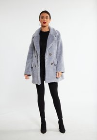 Grå fluffy dobbeltknappet frakke med knapdetaljer og store revers, parret med sorte leggings og ankelstøvler. Glat tekstur.