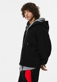 Černá prošívaná bunda s kapucí, předním zipem a předními kapsami; vrstvena přes šedou kostkovanou košili; texturovaná tkanina; volný střih.