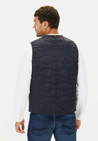 Marineblauer, gesteppten Weste mit Wellenmuster, aus glänzendem Stoff, mit runder Kragen, die über einem weißen Langarmshirt getragen wird.