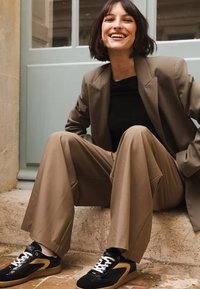 Beige oversized blazer över en svart topp, matchad med lösa bruna byxor och svarta sneakers med gulddetaljer, sittande på en stenyta.