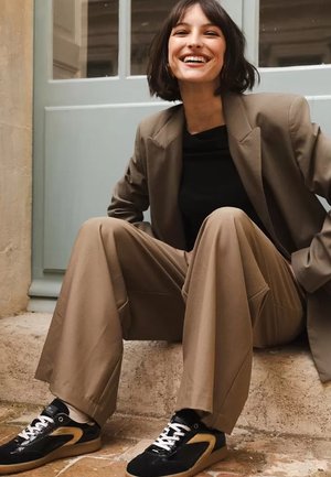 Blazer beige de gran tamaño sobre una blusa negra, combinado con pantalones holgados color canela y zapatillas negras con detalles dorados, sentado sobre una superficie de piedra.