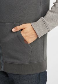 Gray sweatshirt with a smooth texture, featuring a front pocket and contrasting gray sleeves. Includes a small branded tag on the pocket.