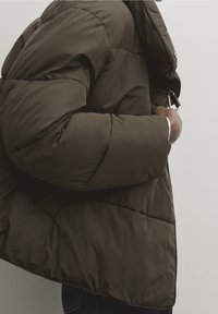 Donkergroene pufferjack met een gewatteerd ontwerp, hoge kraag, ritsluiting en metalen hardwaredetails. Zachte, gladde textuur.