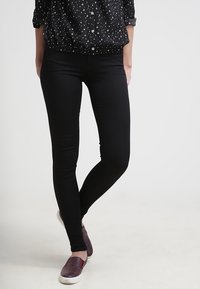 Osoba v černých slim-fit kalhotách, černé košili s bílými hvězdami a tmavě červených slip-on botách, stojící s překříženýma nohama.