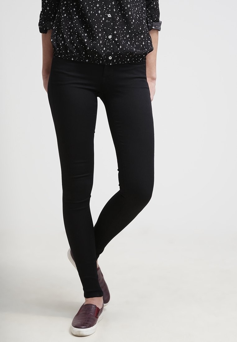 Osoba v černých slim-fit kalhotách, černé košili s bílými hvězdami a tmavě červených slip-on botách, stojící s překříženýma nohama.