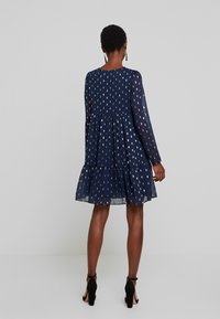 Kobaltblauwe doorschijnende jurk met metallic bladpatronen, lange mouwen en een geplooide rok. Heeft een ronde halslijn en een vloeiende silhouet.