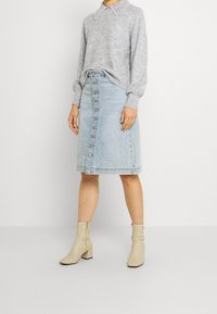 Femme en pull gris avec col perlé, jupe en denim bleu clair boutonnée sur le devant et bottines beige, debout devant un fond blanc.