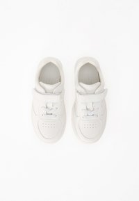 Une paire de baskets blanches pour enfants avec des bandes Velcro et des lacets sur un fond blanc, vue de dessus.