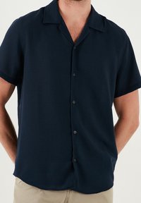 Granatowa koszula na krótki rękaw z guzikiem i kołnierzykiem, wykonana z gładkiego materiału o luźnym kroju i subtelnej fakturze.