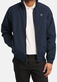 Marinblå lätt bomberjacka med dragkedja fram och elastiska ärmslut, bärs över en vit polo-skjorta. Har logotyp på bröstet.