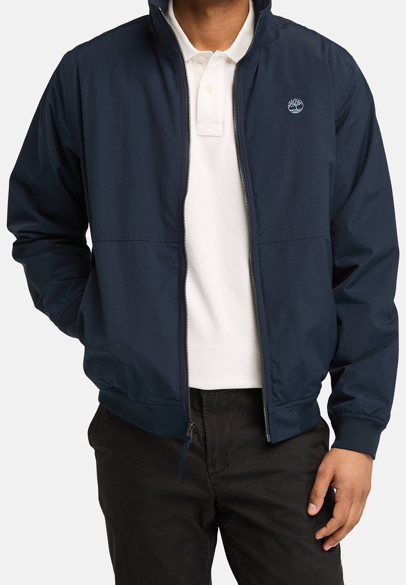 Marinblå lätt bomberjacka med dragkedja fram och elastiska ärmslut, bärs över en vit polo-skjorta. Har logotyp på bröstet.