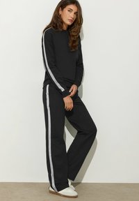 Schwarzes Sweatshirt- und Hosen-Set mit silbernen Seitestreifen. Hergestellt aus weichem Stoff, mit einer lockeren Passform und gerippten Bündchen. Weiße Sneaker runden den Look ab.