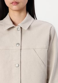 Chemise en denim beige à rayures, avec un col pointu, des boutons-pression argentés et une poche poitrine. Le tissu a une texture douce et des coutures visibles.