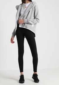 Ljusgrå zip-up hoodie med vita detaljer, bärs över en vit t-shirt och svarta leggings, ihop med svarta sneakers.