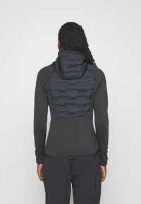 Sort jakke med en tætsiddende design, tekstureret mørkt stof på bagsiden og hætte. Har lange ærmer og en glat, strømlinet silhuet.