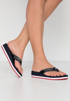 Navy flip-flop sandals with a textured strap featuring the brand "TOMMY" in white, and a wedge heel with a red and white accent.